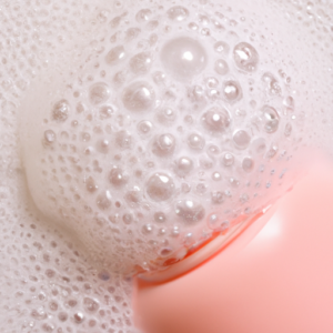 A close-up of a pink facial cleanser bottle with bubbles and foaming suds.